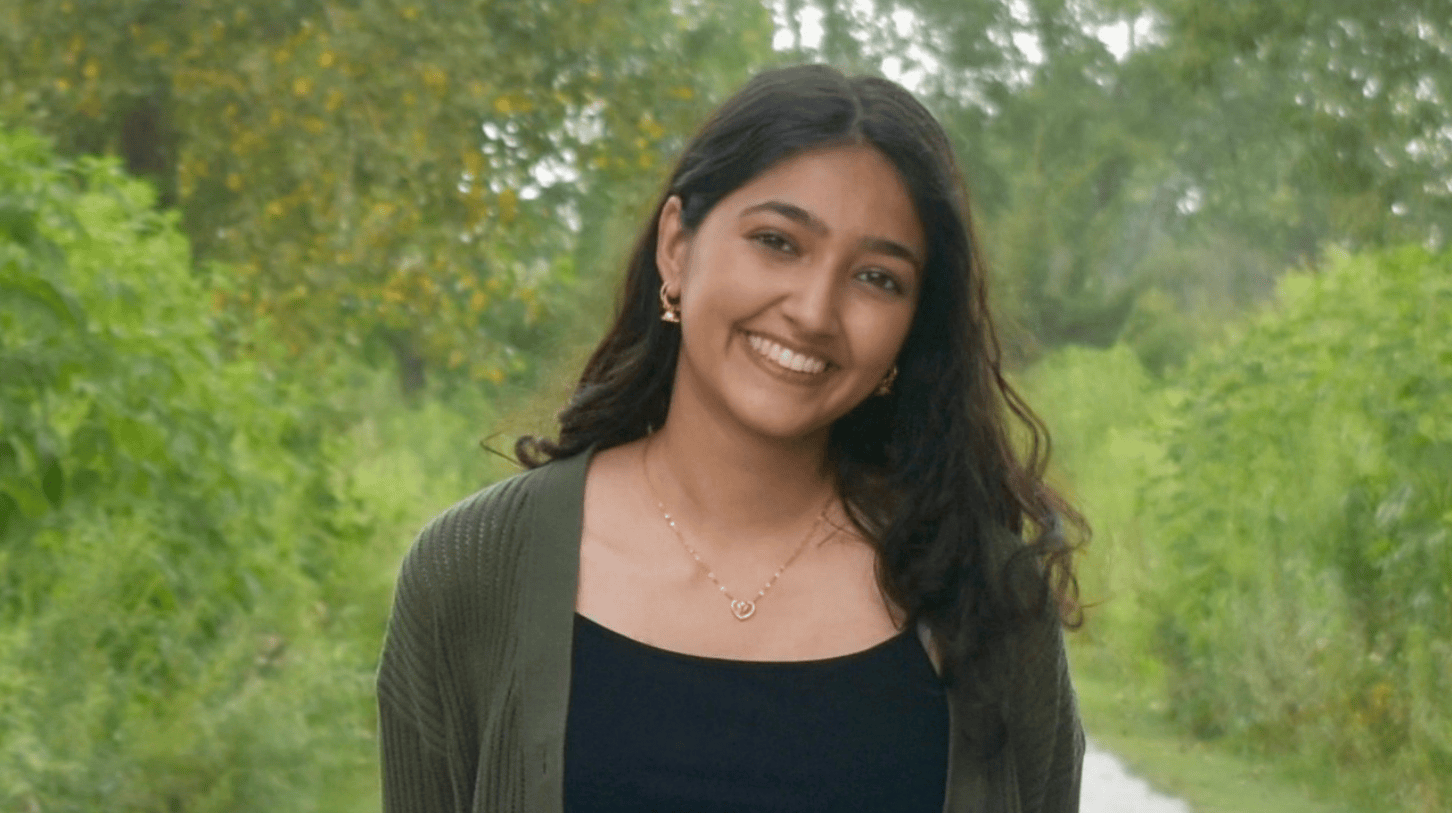 Portrait of a current Gannon Scholar with long dark hair, smiling, wearing a black top and green cardigan with a heart-shaped necklace, standing on a tree-lined path with greenery in the background.